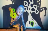 A wooden puppet with large eyes sits on a wooden shelf. It is holding a green leaf shaped like a heart in its left hand. There is an illustration of a white tree against a dark blue wall behind the puppet